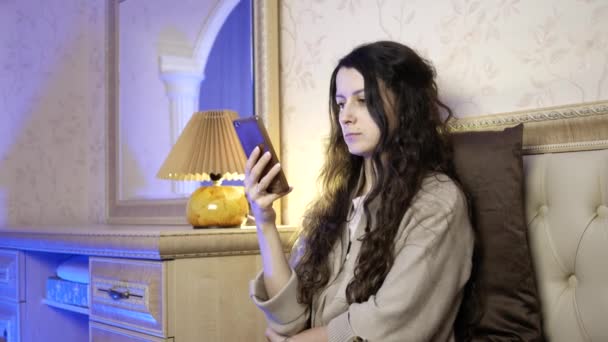 Красивая молодая женщина использует мобильный телефон, сидя на кровати в спальне — стоковое видео