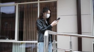 Coronavirus koruması. Tıbbi koruyucu maskeli genç bir kadın şehrin caddesinde akıllı telefonuyla dikiliyor.