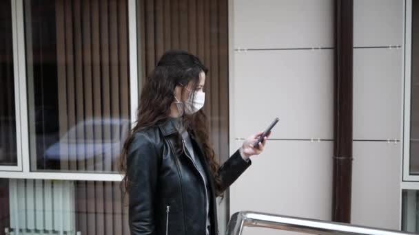 Vista lateral da mulher caucasiana com cabelos escuros para fora e sobre nas ruas da cidade durante o dia, usando máscara facial contra a poluição do ar e Coronavirus Covid-19, usando smartphone — Vídeo de Stock