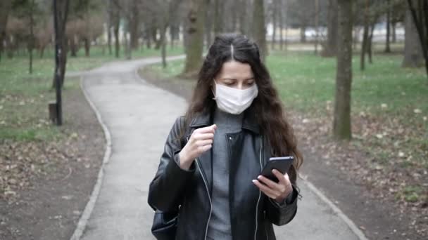 Maskeli genç kadın park ve cadde boyunca yürür telefon kullanır. Viral mikropların yayılmasını önlüyor. Pandemik koronavirüs — Stok video