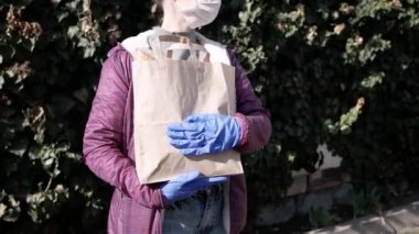 Yiyecek teslimatı yapan koruyucu maskeli genç bir kadın ve eldivenli bir çanta ve mağazadan alınmış ürünler taşıyor. salgın