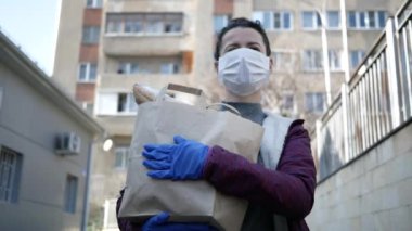 Yiyecek teslimatı yapan koruyucu maskeli genç bir kadın ve eldivenli bir çanta ve mağazadan alınmış ürünler taşıyor. salgın