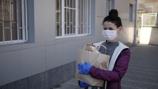 Доставка їжі молода жінка в захисній масці і рукавичках несе сумку з продуктами з магазину. пандемія — стокове відео