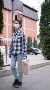 Lateks eldivenli ve tıbbi maskeli genç bir kadın karantina döneminde müşterilerine ürünler ve mallarla birlikte paket teslim ediyor. Yemek. Dikey video