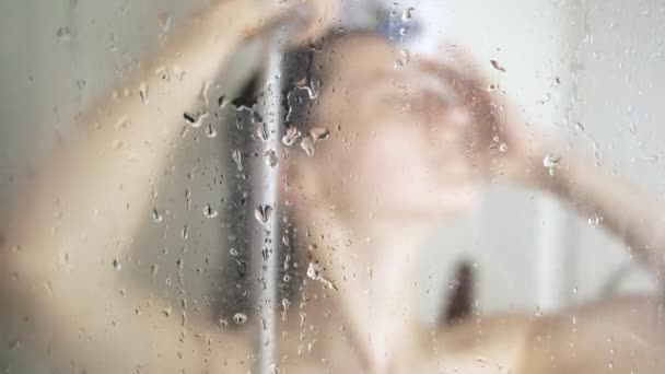 Жінка приймає душ у ванній, миючи тіло під душем. За склянкою в душі. Вибірковий фокус на склі — стокове відео