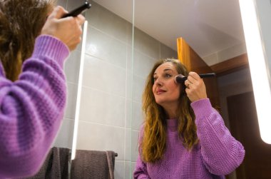 40 yaşında kadın makyaj yapıyor ve işe gitmeden önce banyoda cildine bakıyor.