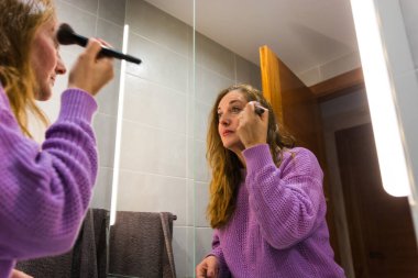 40 yaşında kadın makyaj yapıyor ve işe gitmeden önce banyoda cildine bakıyor.