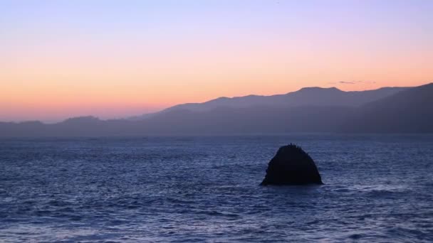 Imágenes relajantes de un sol poniente detrás del horizonte de montañas sobre el océano con aves marinas voladoras alrededor de las rocas. Hora dorada del océano — Vídeo de stock