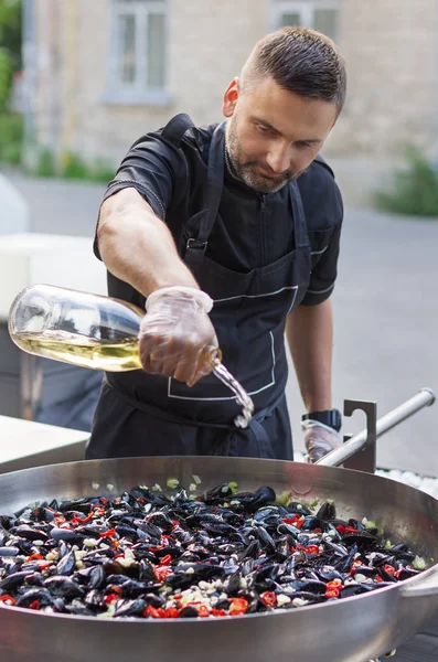 Hombre, está preparando mejillones en una sartén grande en la calle, vierte vino blanco . — Foto de Stock