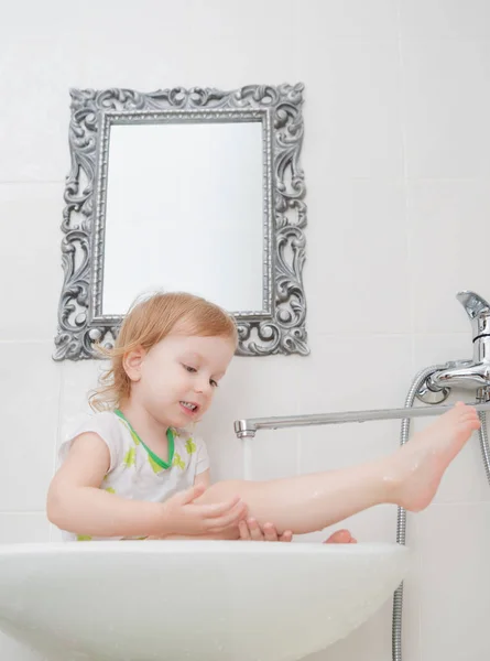 Pequeña niña divertida sentada en el lavabo y lava su pierna, jugando con gotas de agua y salpicaduras . —  Fotos de Stock