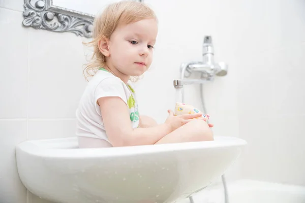 Graciosa niñita sentada en el lavabo y se preguntaba bajo el agua corriente y salpicaduras . —  Fotos de Stock