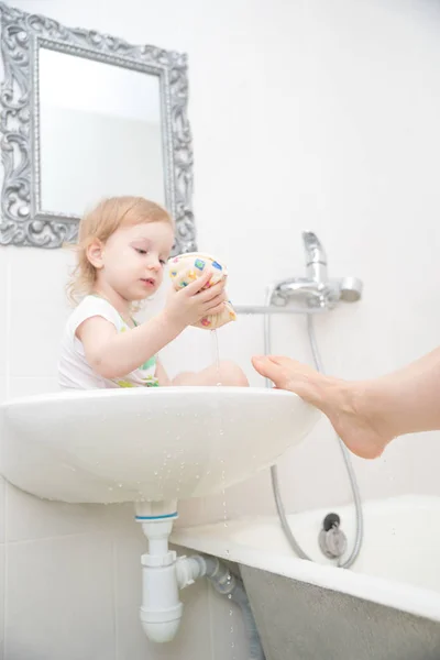 Pequeña niña divertida sentada en el lavabo y lava su pierna, jugando con gotas de agua y salpicaduras . —  Fotos de Stock