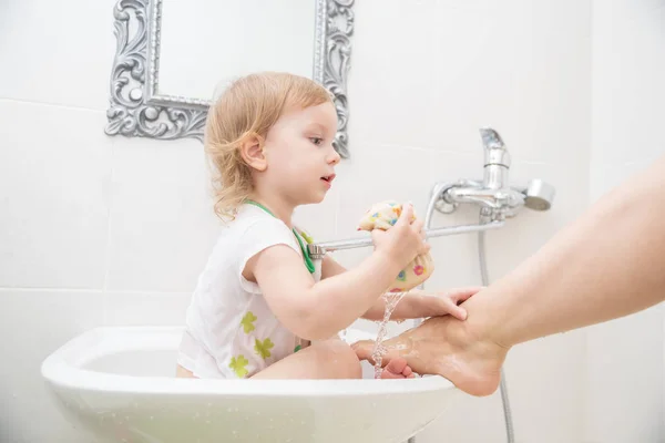 Pequeña niña divertida sentada en el lavabo y lava su pierna, jugando con gotas de agua y salpicaduras . —  Fotos de Stock