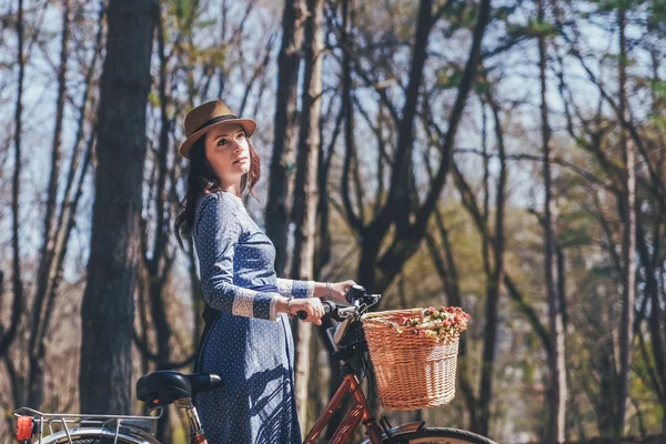 Mulher sorridente jovem bonita com cabelo escuro curto e chapéu de pé perto de bicicleta com cesta de enorme buquê de camomilas.Menina de verão desfrutando da natureza ao ar livre.Fundo verde . — Fotografia de Stock