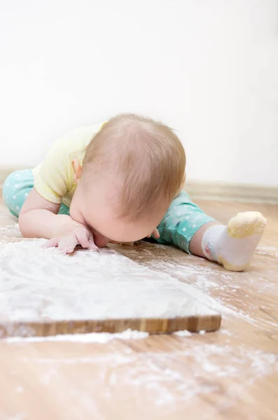 Una niña linda con en la cabeza está sentada en el piso de la cocina, huele la harina, está muy manchada con harina de trigo —  Fotos de Stock