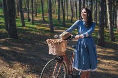 Kısa siyah saçlı ve şapka ayakta yakınındaki büyük chamomiles buket sepet ile bisiklet ile güzel genç gülümseyen kadın. Açık doğanın keyfini yaz kız. Yeşil arka plan.