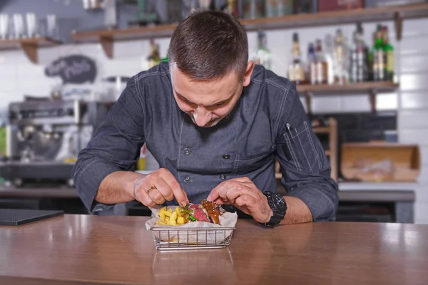 Chef alegre en uniforme, poniendo una deliciosa hamburguesa con papas fritas en la cesta . — Foto de Stock