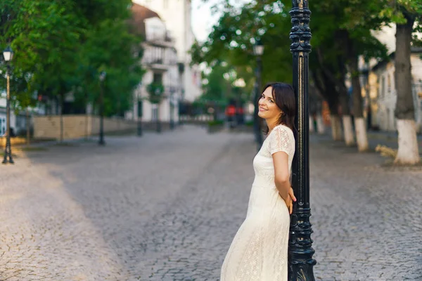 Mulher bonita nova, dark-haired, está com suas costas contra o fonor na rua no centro da cidade em um dia ensolarado, em um vestido branco com uma rua no fundo. Retrato exterior . — Fotografia de Stock