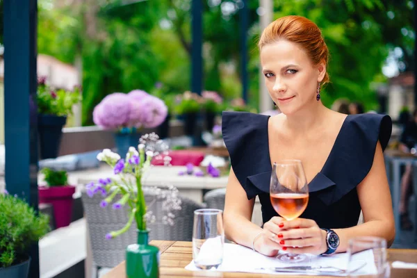 Parlak kırmızı saçlı güzel bir akıllı kadın portresi kapatın. Siyah elbiseli şık genç kız bir kadeh şarap ile bir şehir Restoran terasında eller.