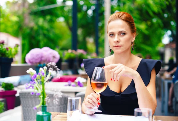 Parlak kırmızı saçlı güzel bir akıllı kadın portresi kapatın. Siyah elbiseli şık genç kız bir kadeh şarap ile bir şehir Restoran terasında eller.