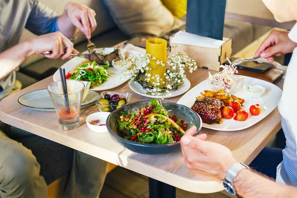Am Tisch essen zwei Männer zu Abend, essen ein Steak, mit einem Salat auf einem weißen Teller, mit Gabel und Messer in der Hand. Stockbild