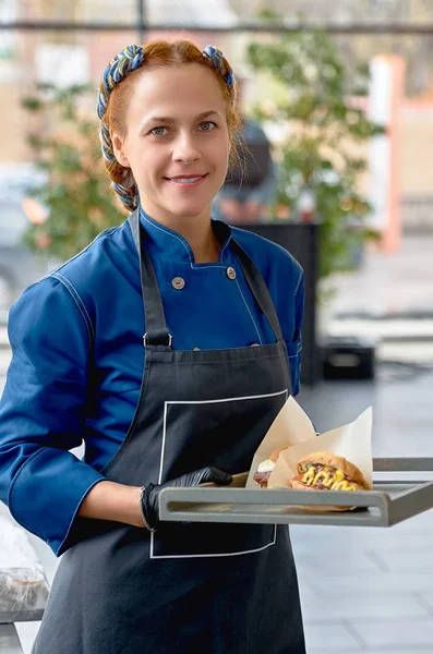 Cheerful, young, chef in apron, presenting a tray with a hamburg — Stock Photo, Image