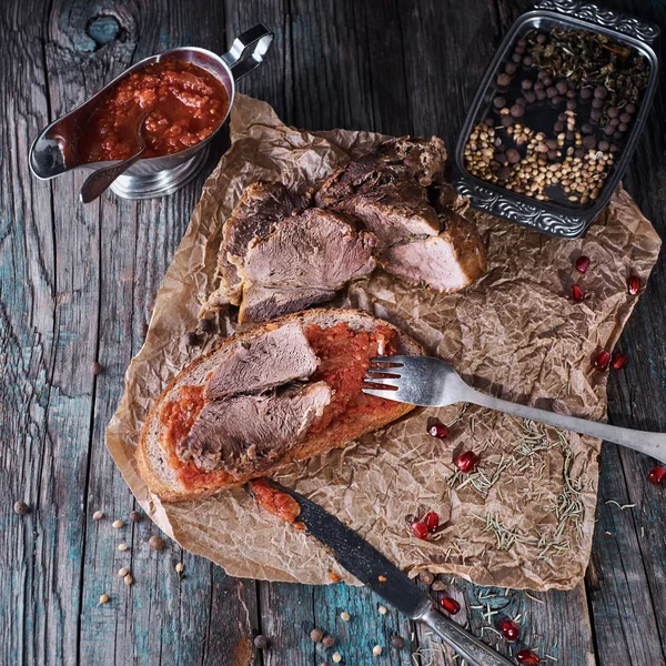 Carne de porc suculentă coaptă, bucăți feliate pe hârtie pergament, aproape cu pâine și sos de roșii, fundal rustic din lemn închis — Fotografie, imagine de stoc