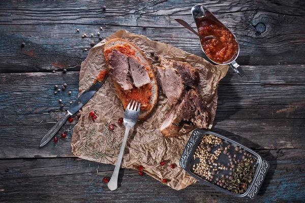 Carne de cerdo suculenta al horno, trozos cortados en papel pergamino, primer plano con pan y salsa de tomate, fondo rústico de madera oscura — Foto de Stock