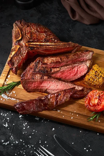 Cerrar filete de ternera a la parrilla medio raro rebanado con maíz y tomate, pimienta y romero en la tabla de cortar la carne sobre fondo de madera oscura — Foto de Stock