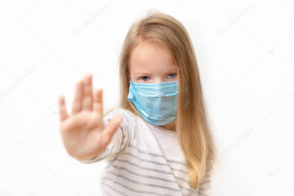 little girl putting up her hand, to say no, or stop, with a serious frowned expression in a medical mask on his face on a light background, close-up.