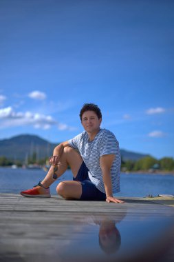 Yakışıklı adam güneş gözlüğü takıp iskelede oturuyor ve kameraya bakıyor..