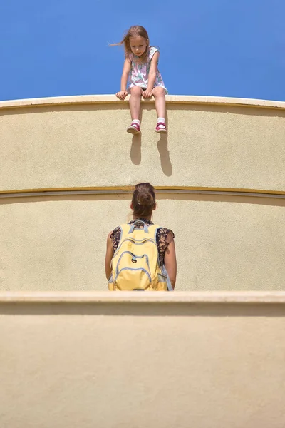 Little girl sits on the edge of the wall, a stone wall her legs dangling in the air and mom reaches for her — Stock Photo, Image