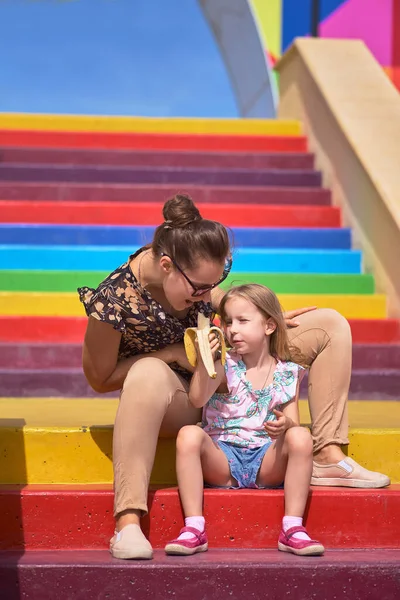 Madre joven con gafas y una hija están sentados en una escalera de colores. Concepto de Protección Infantil, Día de las Madres — Foto de Stock