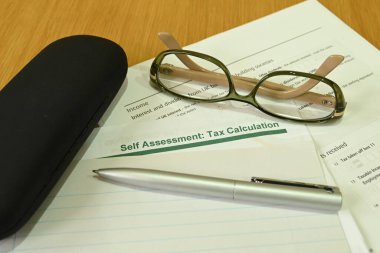 Close up view of a self assessment tax return form with a pen, a pair of glasses and a glasses case on a wooden table clipart