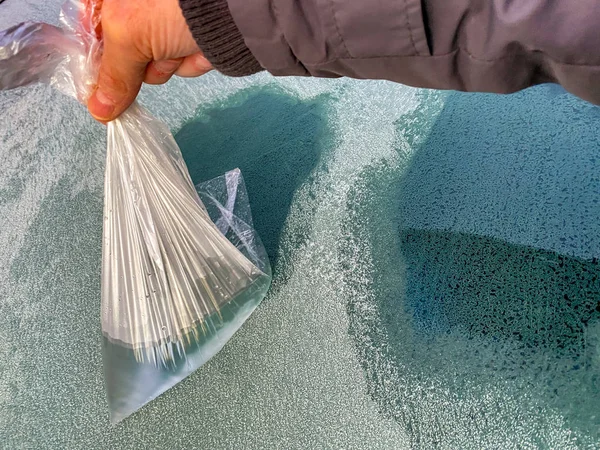 Person using a plastic bag of very hot water to melt ice crystals from heavy frost on the windscreen of a car on a winter morning.