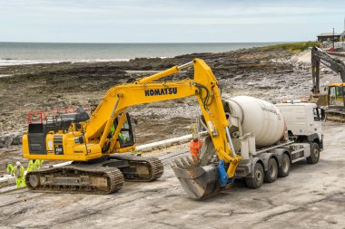 PORTHCAWL, WALES - Haziran 2018: Porthcawl 'daki deniz önü ve deniz savunmasını güçlendirme amaçlı bir projede kullanmak üzere ağır bir kazıcının kovasına karışık beton boşaltan beton karıştırıcı.