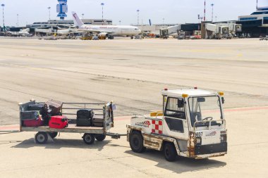 Milano Malpensa Aiport 'ta önlüğe park edilmiş küçük bir traktör ve bagaj vagonu. Arka planda bir uçak var..