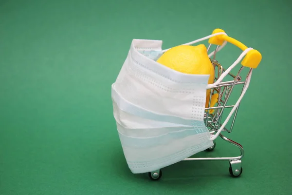 Uma máscara médica é colocada em um carrinho de compras contendo um limão fresco. Fundo verde . — Fotografia de Stock