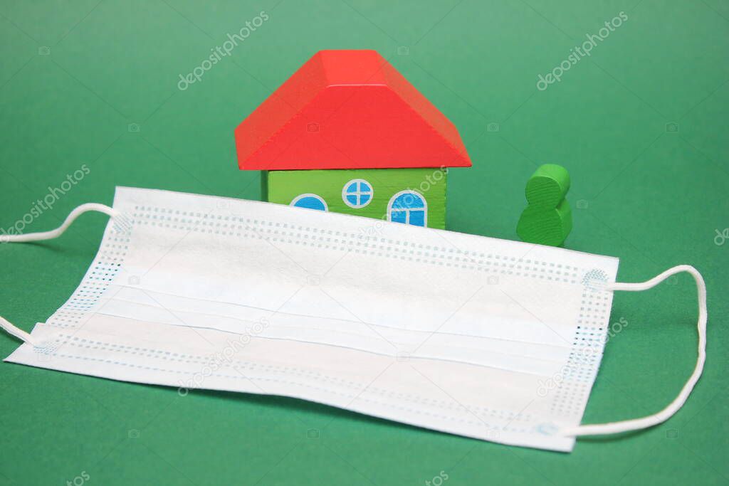 Medical mask near wooden figures of a house and a person. Concept of Isolation and home treatment during the coronavirus pandemic. Green background.