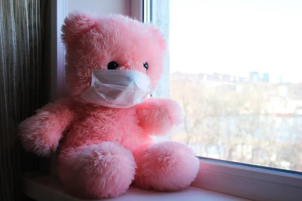 Un oso rosa de juguete en una máscara médica se sienta en el alféizar de la ventana cerca de la ventana. Cuarentena. pandemia de Coronavirus . —  Fotos de Stock