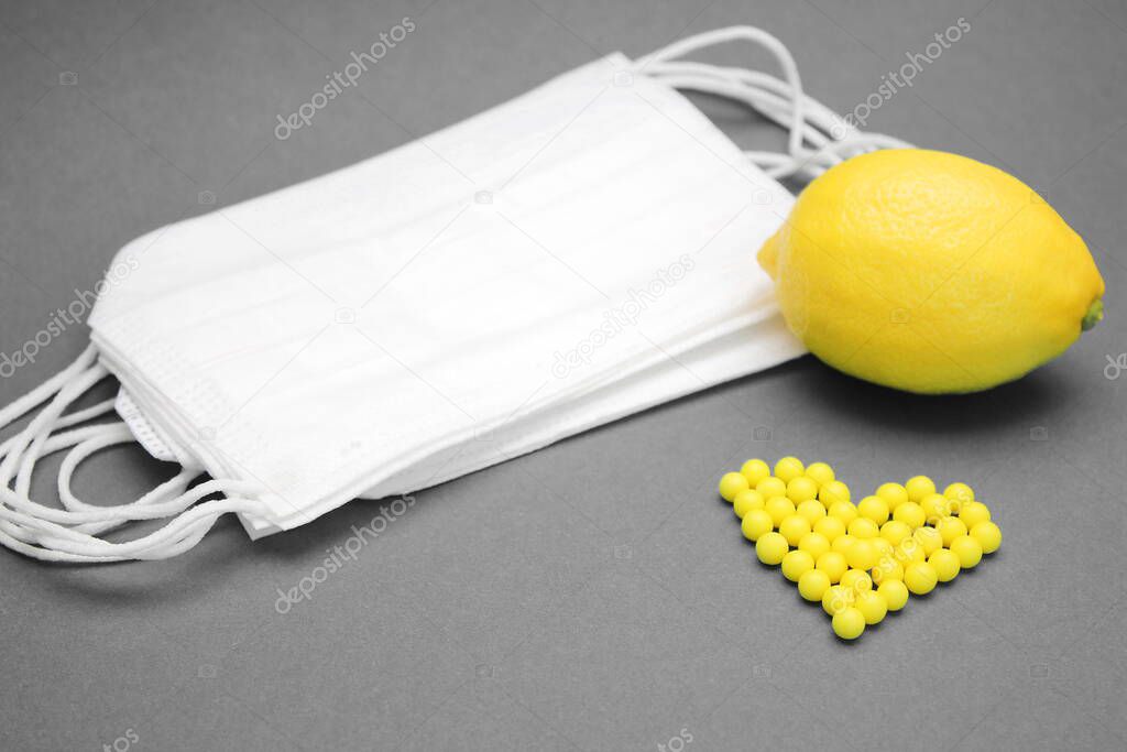 Lemon next to medical masks and ascorbic acid vitamins are laid out in the shape of a heart on a green background.