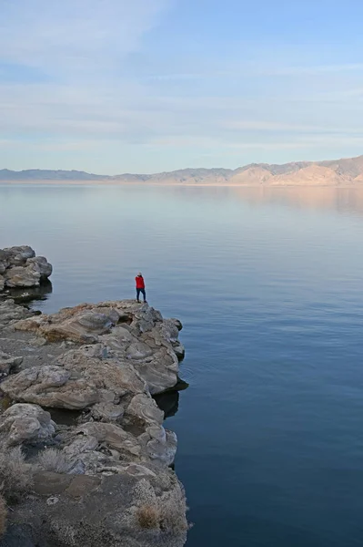 Wanita muda bersweater merah berdiri di atas batu oleh Pyramid Lake, Nevada pada sore hari . — Stok Foto