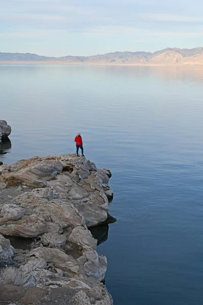 Wanita muda bersweater merah berdiri di atas batu oleh Pyramid Lake, Nevada pada sore hari . — Stok Foto