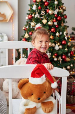 Noel bebeği. Beyaz yatakta, çocuk odasında, Noel ağacının arka planında gülümseyen küçük kız. Çocuk odasında Noel. Komik bebek yalan söylüyor. Sevimli küçük bebek.