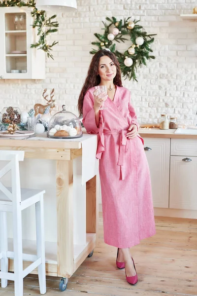 Woman at home for christmas. Woman preparing at home. Smiling woman in pink dress in kitchen. White kitchen interior. Modern kitchen with table and kitchenware. Kitchen in luxury home.