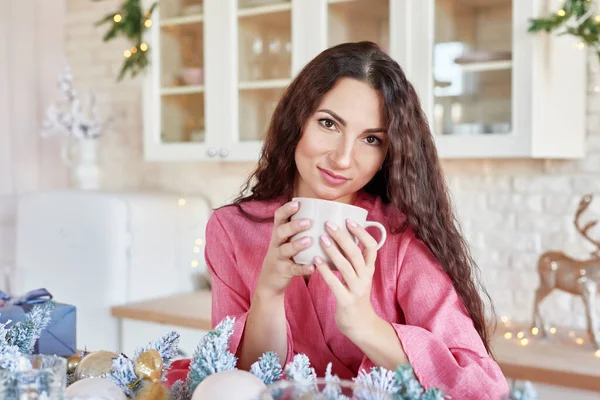 Boldog fiatal nő rózsaszín ruha csésze karácsonyi díszített konyha. gyönyörű barna csésze kakaó a konyhában, karácsonyi dekoráció. Világos fehér konyha belseje karácsonyra. Asszony otthon. — Stock Fotó