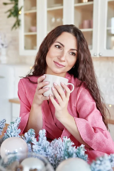 Šťastná mladá žena v růžových šatech s šálkem v vánoční zdobené kuchyni. krásná brunetka s šálkem kakaa v kuchyni s vánoční výzdobou. Světle bílý kuchyňský interiér na Vánoce. Žena doma — Stock fotografie