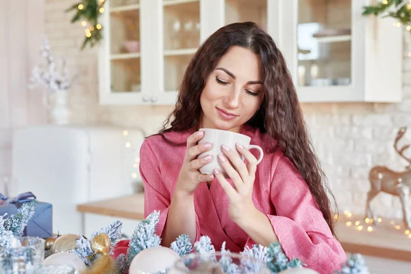 Joyeux jeune femme en robe rose avec tasse dans la cuisine décorée de Noël. belle brune avec tasse de cacao dans la cuisine avec décor de Noël. Intérieur de cuisine blanc clair pour Noël. Femme à la maison — Photo