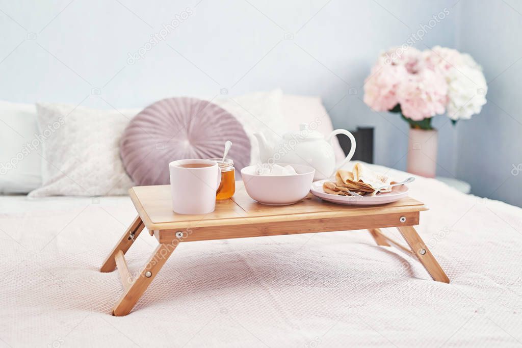 Breakfast in bed in hotel room. Accommodation. Breakfast in bed with tea cup with pancakes on tray on bed background top view. Copy Space. Romantic valentine's day breakfast. Cozy breakfast Maslenitsa