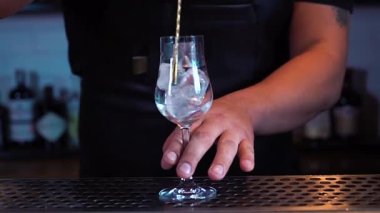 Barmen kokteyl bardağında buz karıştırıyor. Birisi barda kokteyl hazırlıyor. Buz camda dönüyor..